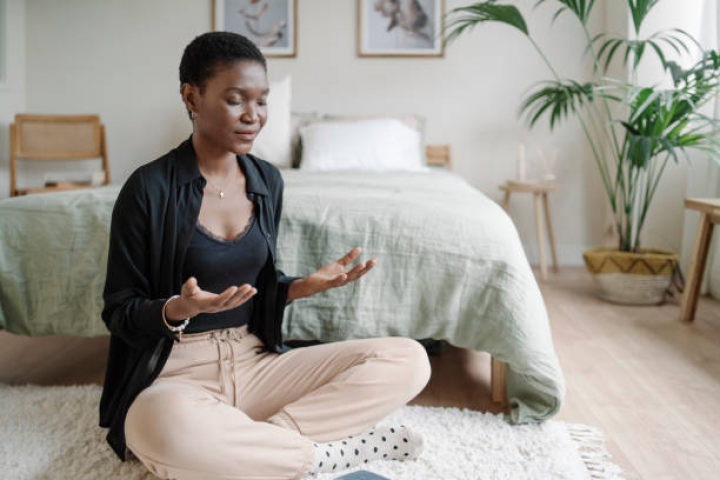 alt="alt="Relaxed black woman sitting on the floor practicing meditation after reading book at home.""