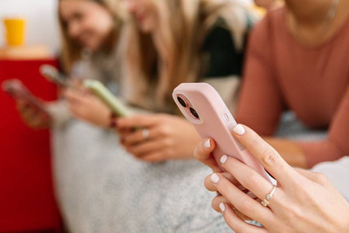 alt="alt="Close up view of young adult women lying on the bed using their smartphones. Gen Z friends relaxing together with technology. Digital lifestyle and friendship concept.""