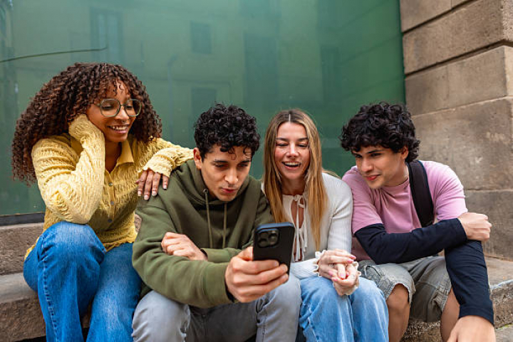 alt="alt="Diverse group of young adults sitting on urban steps, smiling and sharing a smartphone while enjoying a casual, connected moment of friendship, laughter and togetherness""