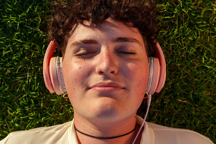 alt="alt="Young transgender woman lying on grass with closed eyes, enjoying music with headphones in the park at sunset""
