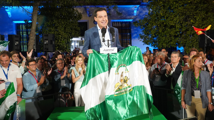 Subido en un escenario, rodeado de simpatizantes del PP, Juanma Moreno Bonilla sostiene una bandera de Andalucía