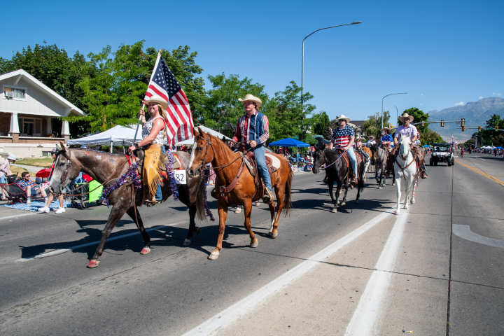 alt="alt="Artistas ecuestres con sombreros de vaquero desfilan a caballo por la calle portando una bandera estadounidense durante el Gran Desfile del Festival de la Libertad de Estados Unidos en Provo, Utah.""