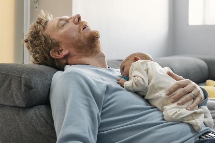 Un padre exhausto duerme en el sofá, sosteniendo a su bebé.
