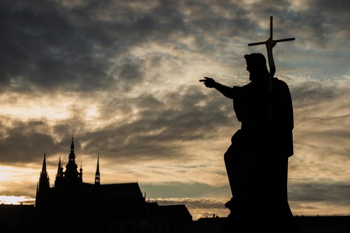 Silueta de la estatua de San Juan Bautista en el Puente de Carlos en Praga, con la Catedral de San Vito al fondo, al atardecer.