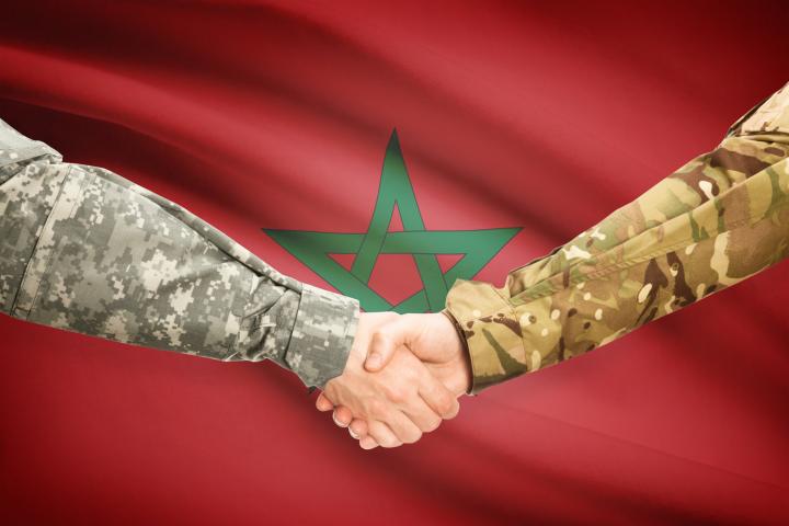 Ilustración digital de dos hombres uniformados estrechándose la mano ante la bandera de Marruecos.