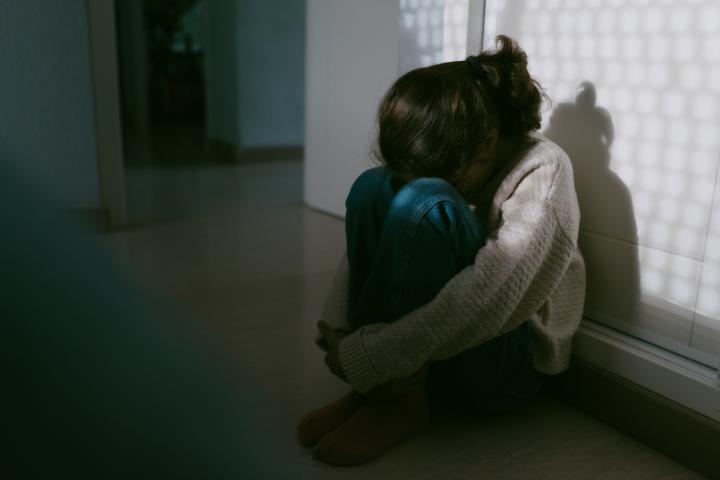 Una joven sentada en el suelo de su habitación con la cabeza apoyada en las rodillas.