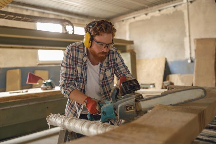 Carpintero con gafas cortando madera con motosierra en un taller.