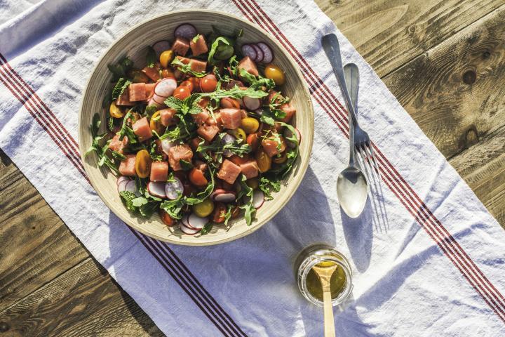 Una colorida y fresca ensaladera con cubos de sandía, tomates cherry, rúcula, rábanos, menta y ralladura de limón. Fotografía tomada bajo el sol radiante en una mesa de picnic en Portugal, durante la primavera. Un pequeño frasco de aderezo, zumo de limón y aceite de oliva acompaña esta ensalada fresca y saludable, ¡lista para comer!