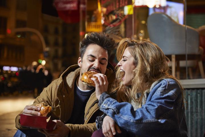 Una mujer alegre le da de comer una hamburguesa a un hombre. Un grupo de amigos, un hombre y una mujer, están sentados en la terraza de un restaurante. Visten ropa informal, por la noche.
