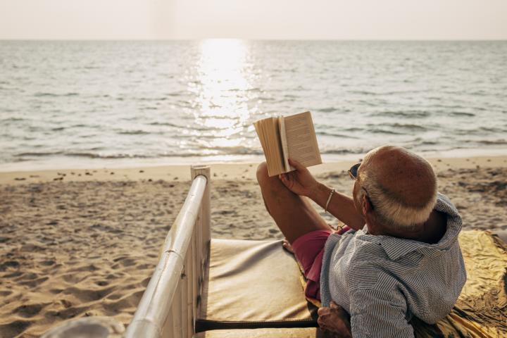 Hombre mayor relajado tumbado en la cama mientras lee un libro en la playa al atardecer.