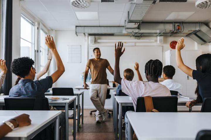 Alumnos levantan la mano en una clase frente a su profesor.