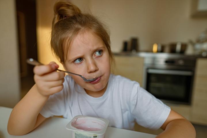 Una chica mona come yogur con cuchara con mucho apetito, tiene la boca sucia, se divierte y hace muecas. Escena cotidiana, en casa, en la cocina, primer plano. Retrato