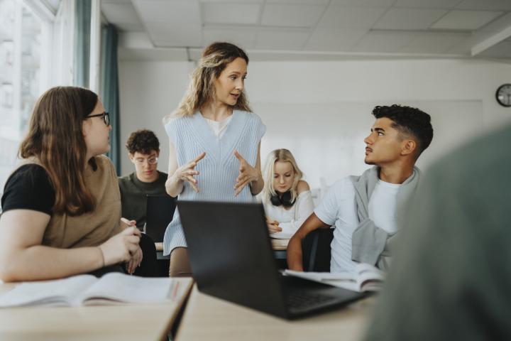 Alumnos atienden en clase a su profesora, quien da una explicación.