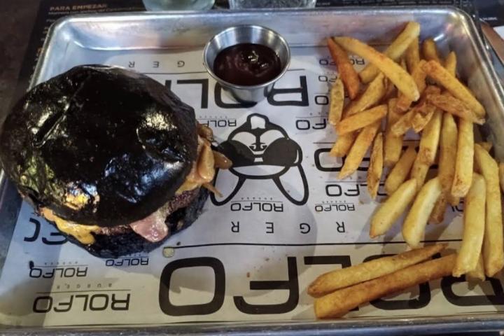 Hamburguesa de pan negro con queso y bacon acompañada de patatas fritas y salsa en bandeja metálica del restaurante Rolfo Tapas & Burger, en Murcia.