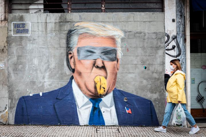 Una mujer con mascarilla lleva a un bebé en brazos mientras pasa junto a un grafiti del artista portugués Kilos. El grafiti satírico mezcla a Donald Trump con un plátano pegado a la pared con cinta adhesiva.