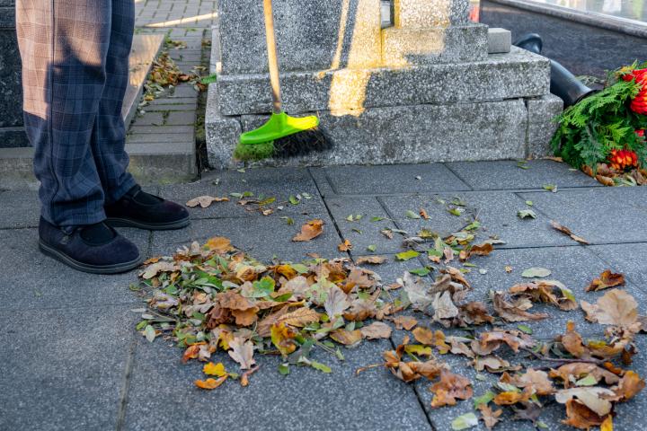 Persona barriendo las hojas de otoño del sendero del cementerio cerca de la tumba, preparándose para el Día de Todos los Santos y cuidando el lugar de descanso conmemorativo de sus seres queridos.