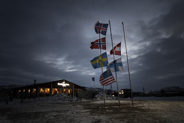Varias banderas de los países nórdicos ondean en el mástil al viento, con el cielo nocturno entre las nubes de fondo. Un paisaje espectacular.
