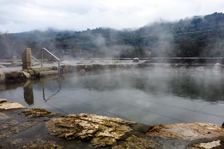 alt="alt="Un manantial termal, con sus aguas tranquilas y cristalinas, se recorta contra el telón de fondo de montañas envueltas en niebla. El vapor se eleva etéreo desde la superficie, sugiriendo la calidez natural de sus aguas que invitan a un baño relajante. Los bordes rústicos de piedra y la barandilla solitaria realzan el encanto intacto de este remanso de paz, invitando a imaginar la apacible sonoridad del silencio, interrumpida únicamente por el suave murmullo del manantial. Testimonio del poder sanador de la naturaleza, esta imagen es una ventana a un momento de calma y renovación.""