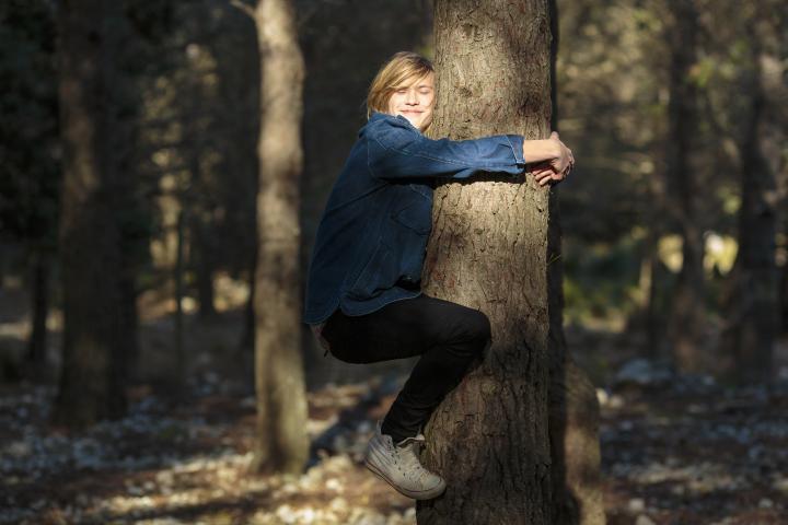 Un niño rubio y ecológico le da un abrazo cariñoso a un árbol en el bosque, de pie en el aire bajo la brillante luz del sol, con los ojos cerrados y luciendo cómodo y sonriente.