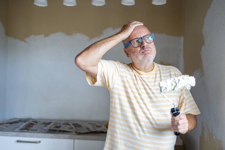 Hombre maduro y cansado con canas sosteniendo el rodillo de pintura mientras está de pie junto a una pared medio pintada