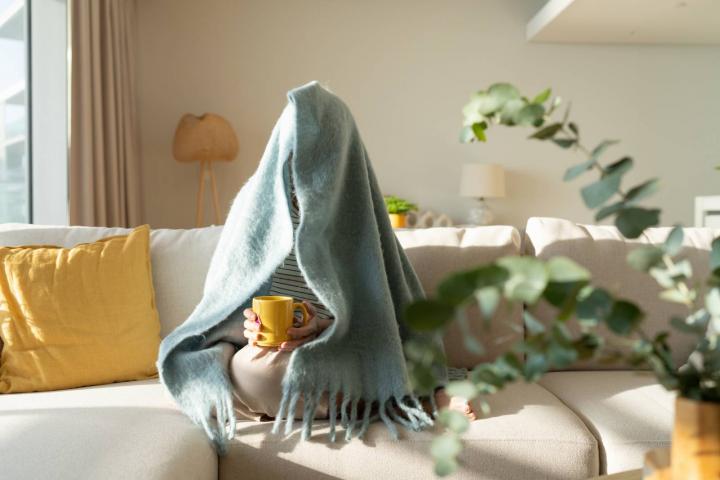 Mujer envuelta en una manta sentada en el sofá y tomando té en el salón