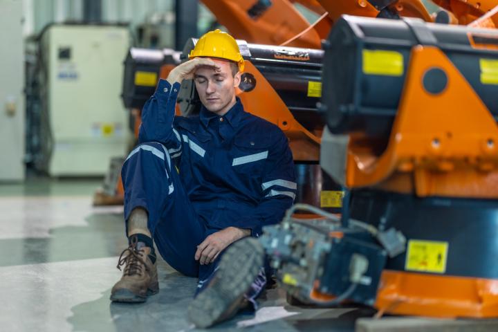 Un técnico de una fábrica se sienta en el suelo de un entorno industrial, con aspecto exhausto y abrumado, en una imagen de archivo.