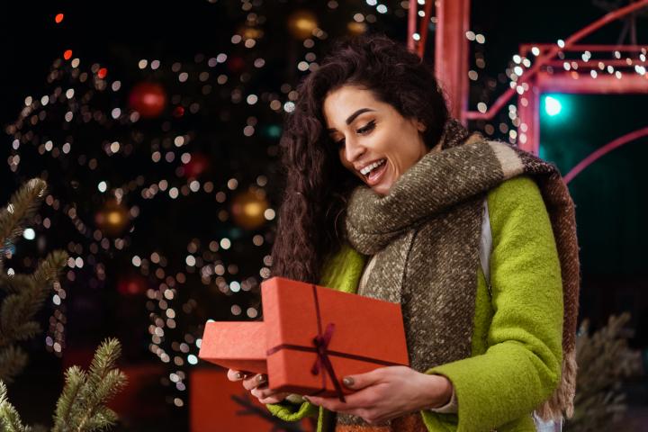 Una mujer abriendo un regalo con cara de sorpresa y un árbol de Navidad de fondo.