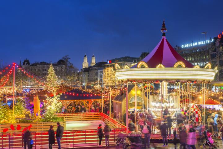 alt="alt="Vista panorámica del mercado navideño de Zúrich en la recientemente reconstruida plaza pública Bellevue / Secheseläutenplatz, justo en frente de la Ópera y al lado del lago de Zúrich, con la emblemática catedral de Grossmunster en la distancia.""