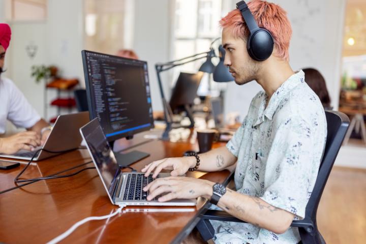 Joven con auriculares trabajando en la computadora en una oficina de startup. Joven profesional de TI trabajando en una oficina de coworking con gente trabajando al fondo.