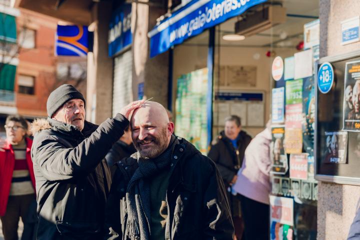 Dos hombres sonríen mientras uno frota un décimo de la Lotería de Navidad en la calva del otro frente a la administración Gaia Loterías en Sant Boi.