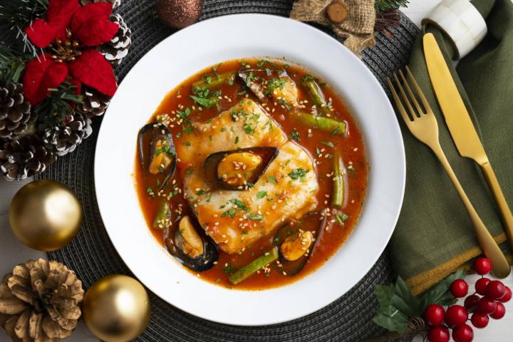 Merluza cocida con tomate y mejillones y presenta en una mesa con decoración navideña