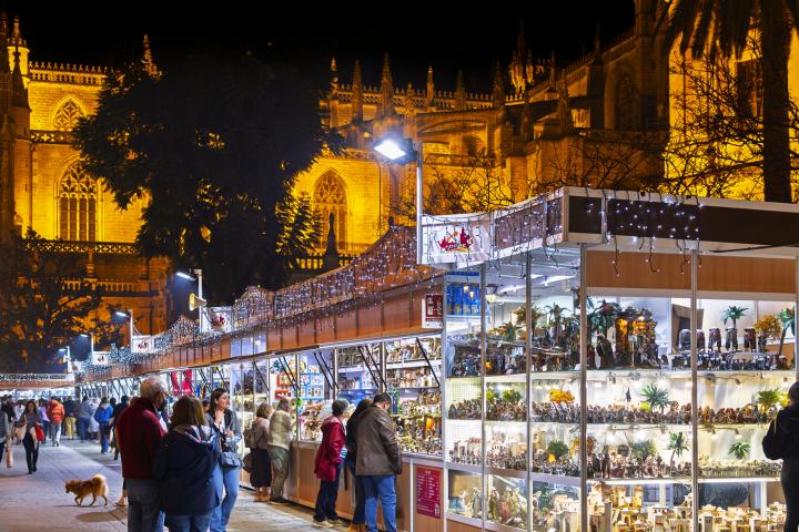 alt="alt="Vista nocturna mientras los españoles visitan el colorido mercado navideño iluminado de la Feria de Belén con decoraciones artesanales junto a la catedral gótica de Sevilla.""