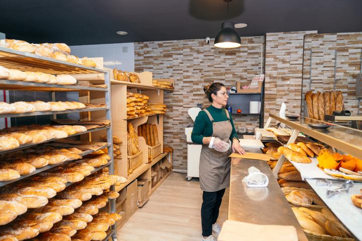 Una trabajadora de panadería entre estantes de panes frescos, manipulando una bolsa de harina en preparación para la siguiente tanda de horneado.
