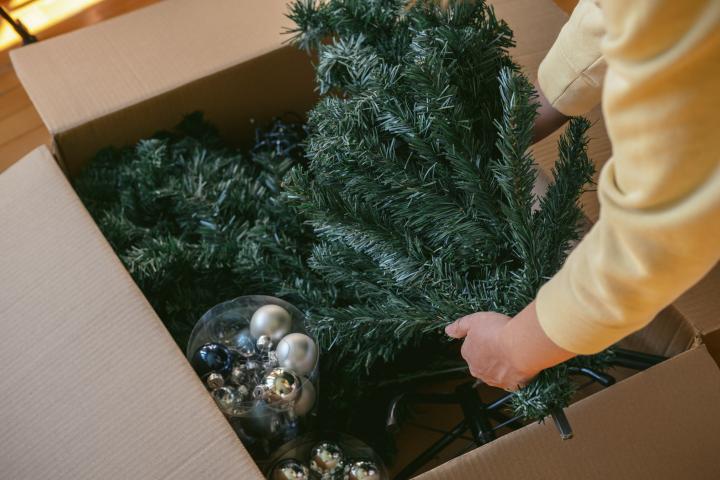 Mano de mujer desempacando un árbol de Navidad artificial, con brillantes bolas plateadas y azules y una guirnalda de una caja. Preparándose para la celebración de Navidad y Año Nuevo.