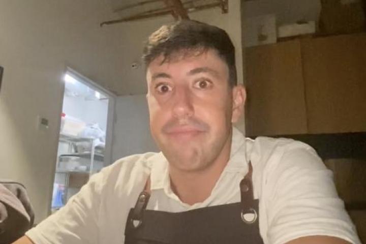 Joven español con delantal, sentado solo en un cuarto del restaurante con mesas y sillas apiladas, durante la Nochebuena en Australia.