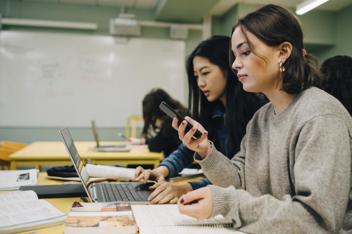 Estudiante de secundaria que usa un teléfono inteligente mientras está sentada junto a un compañero de clase estudiando en una computadora portátil en el aula.