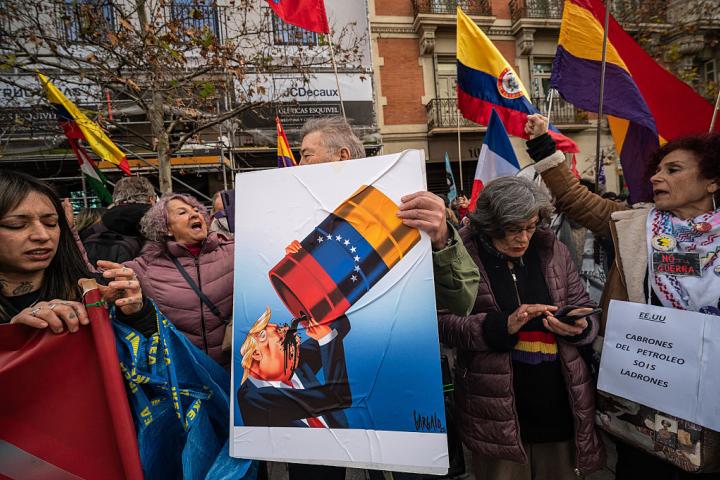 Unos manifestantes ante la embajada de EEUU en Madrid portan un cartel que muestra a Donald Trump bebiendo petróleo venezolano, el 4 de enero de 2026.
