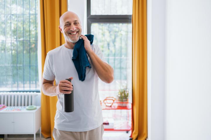 Hombre mayor sonriente después del entrenamiento sostiene una botella y una toalla en un gimnasio en casa brillante
