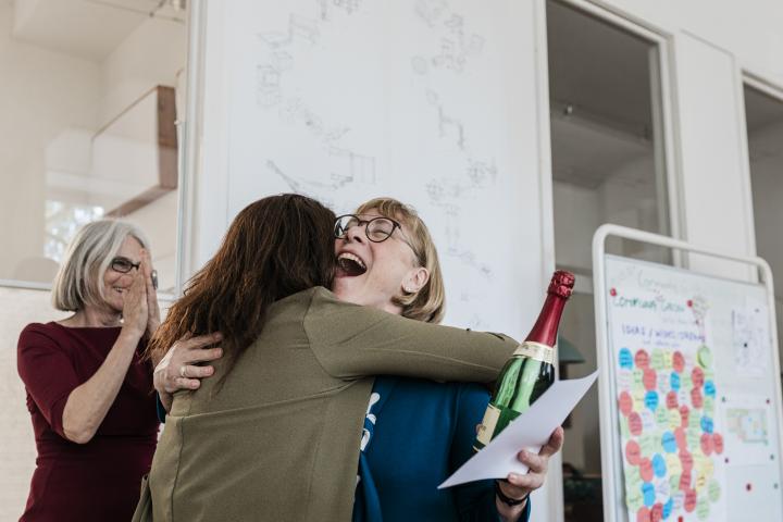 Una mujer mayor sonríe alegremente mientras abraza a un compañero durante una celebración en la oficina. Sostiene una botella de champán y un certificado mientras otro miembro del equipo aplaude de fondo.
