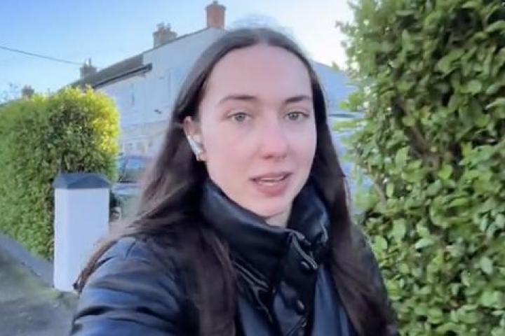 Primer plano de una joven con chaqueta negra acolchada y auriculares caminando por una calle residencial con casas blancas y setos verdes al fondo.