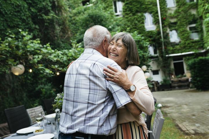 Una pareja de ancianos abrazándose y sonriendo antes de sentarse a hacer una barbacoa con la familia en un patio.