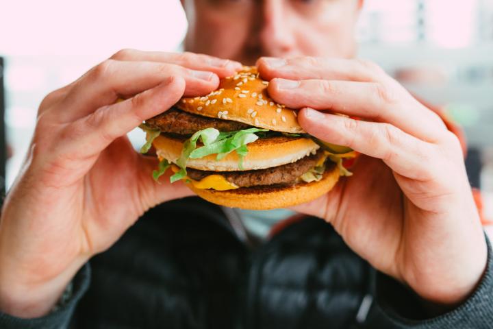 Primer plano a color de un hombre adulto comiendo una hamburguesa recién hecha en un restaurante de comida rápida. La hamburguesa está desordenada, con lechuga y queso derretido sobresaliendo del pan. El hombre tiene unos 30 años y viste ropa informal: una sudadera roja con capucha y un chaleco negro.