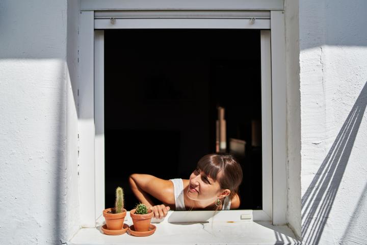alt="alt="Vista frontal de una joven inquietante sacando la cabeza por la ventana y mirando hacia un lado con algunos cactus en la ventana.""