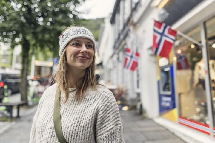 Una mujer con un suéter blanco de punto y una diadema estampada camina por las calles de Bergen, rodeadas de edificios blancos. Sonríe bajo una hilera de banderas noruegas.