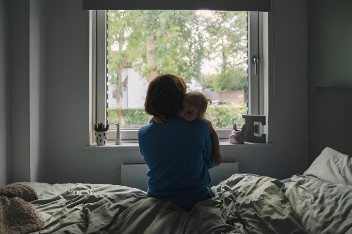 Una mujer sentada en la cama mirando hacia la ventana mientras abraza a su pequeña hija.
