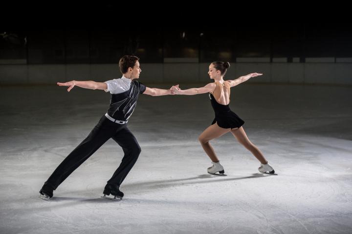 Un hombre y una mujer patinadores artísticos se desempeñan en una pista de hielo, ambos tomados de la mano con el brazo extendido.