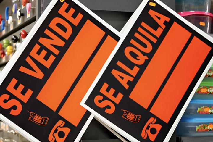 Dos carteles de venta y alquiler en español colgados en un estante dentro de una tienda, con gráficos vibrantes en naranja y negro
