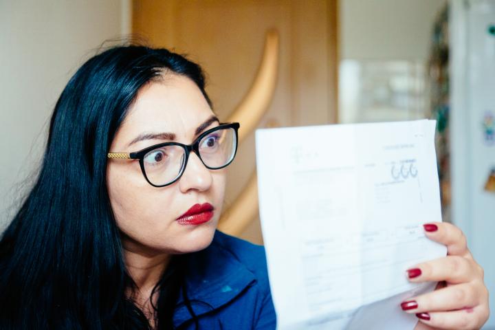 Retrato de una mujer de mediana edad revisando sus facturas de energía en casa, sentada en la cocina. Tiene expresión preocupada y se toca la cara con la mano mientras mira las facturas. El enfoque se centra en la mujer mientras la arquitectura interior del dormitorio está desenfocada.