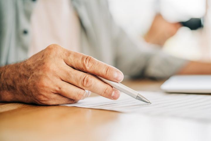 Primer plano de la mano de una persona mayor sosteniendo un bolígrafo, señalando un documento o contrato. El momento de tomar decisiones, como firmar un contrato o revisar documentos.