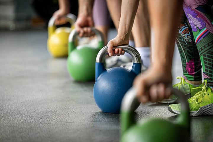 Multiple hands grip kettlebells of different colors, lined up on the floor. The detail shot emphasizes strength, coordination, and functional fitness equipment.